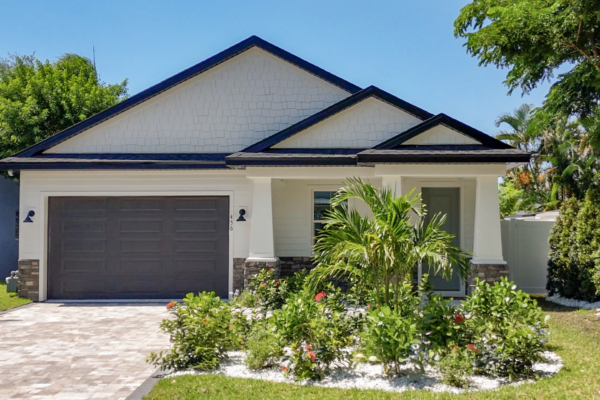 Front view of a modern white house with a gray double garage door, two black-trimmed gabled roofs, and tropical landscaping under a blue sky.