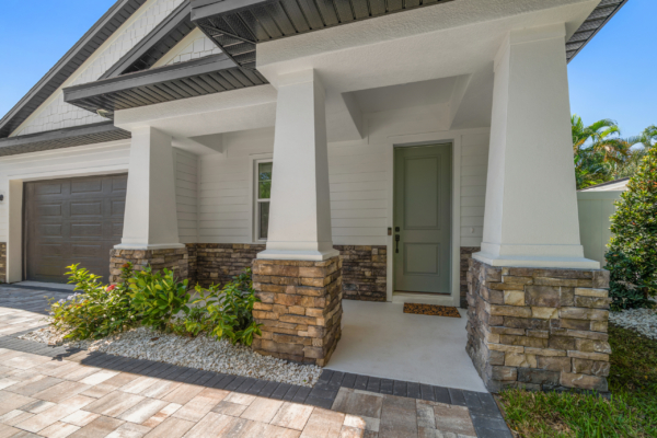 Front view of a modern white house with stone veneer columns, gray garage door, and a green entry door under a covered porch. Bright blue sky.