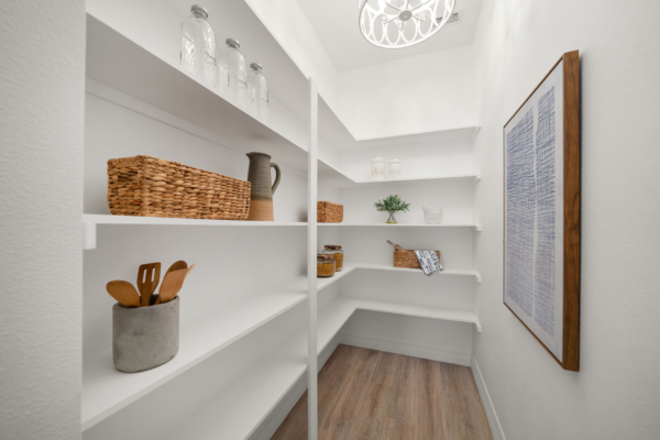 White pantry with open shelves holding wicker baskets, glass jars, and wooden utensils; framed blue print on the right wall; light wood floor and bright, clean space.