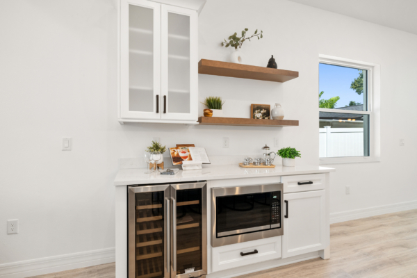 White kitchen wall with built-in glass-front cabinet and two wooden floating shelves displaying plants and decor overlooking a windowed area.