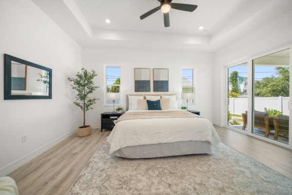 Bright modern bedroom with a large bed, beige bedding, and two nightstands facing glass doors to a patio. A ceiling fan and minimalist art on the wall enhance the space.