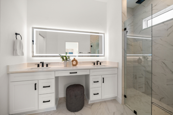 Bright white bathroom vanity with two sinks, black fixtures, a backlit rectangular mirror, and a glass-enclosed marble shower to the right.