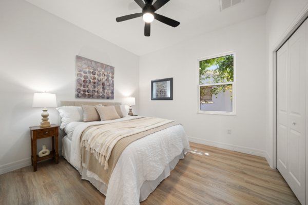 Bright bedroom with white walls, beige bedding, two lamps, round-pattern artwork, a window showing greenery, and a ceiling fan above the bed.
