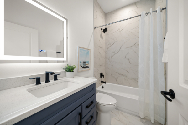 Modern bathroom with a blue vanity, white speckled countertop, and black faucets beneath a backlit mirror with a plant nearby.