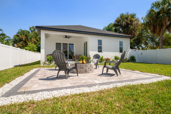 Patio seating area around a fire pit in a sunny backyard with a white house and palm trees.