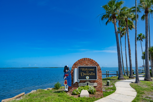 Welcome to Dunedin sign by the waterfront with palm trees and blue sky in the background.