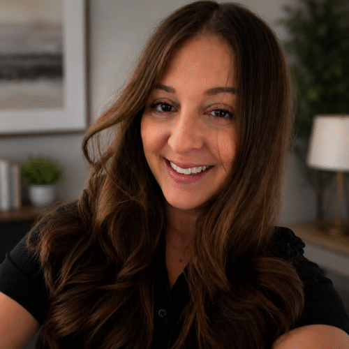 Close-up of a smiling woman with long brown hair wearing a black top, indoors with plants and a lamp in the background.